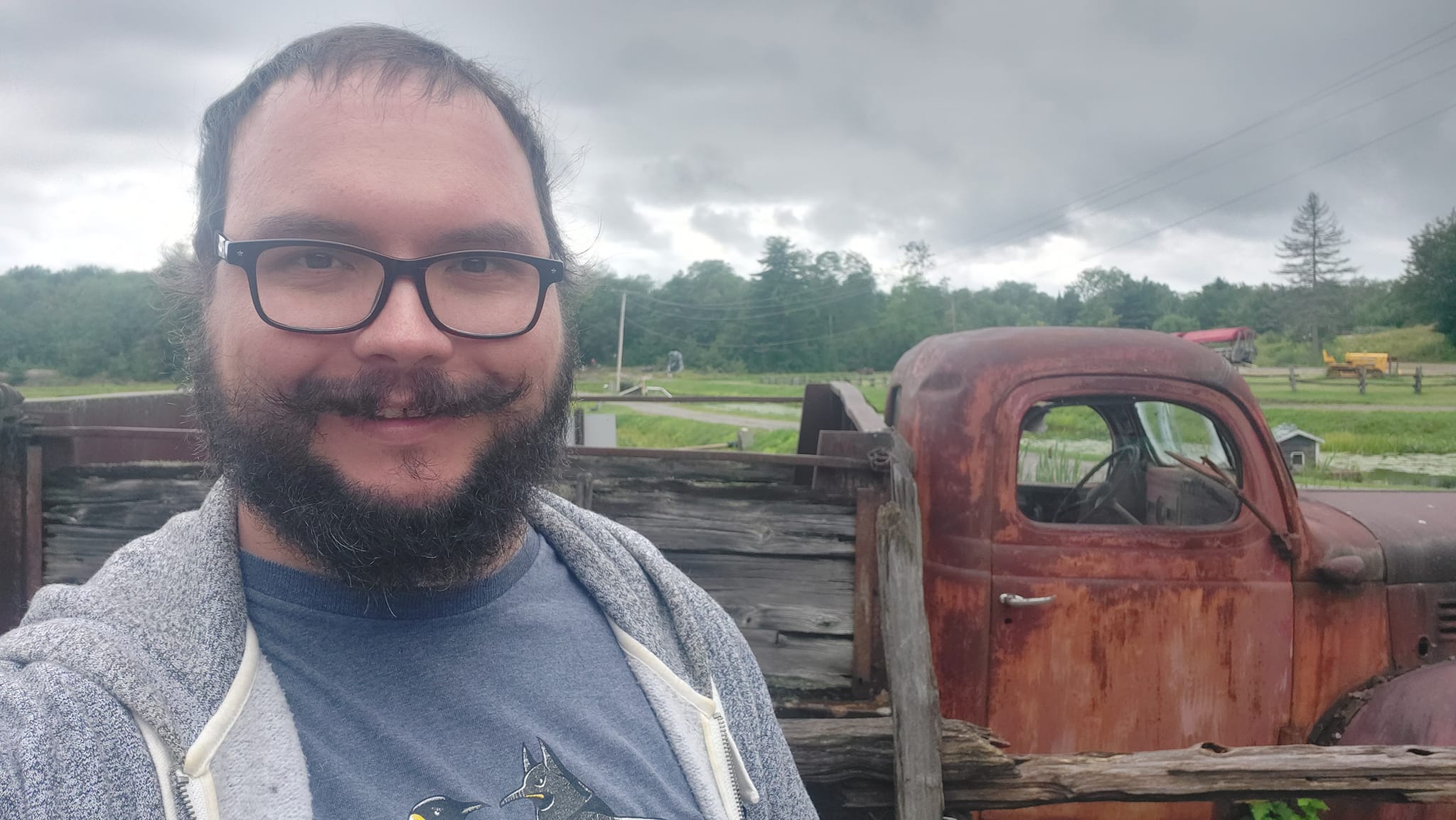 A man with short hair and bushy facial hair stands in front of a rusty old truck, fields appear to be in the background